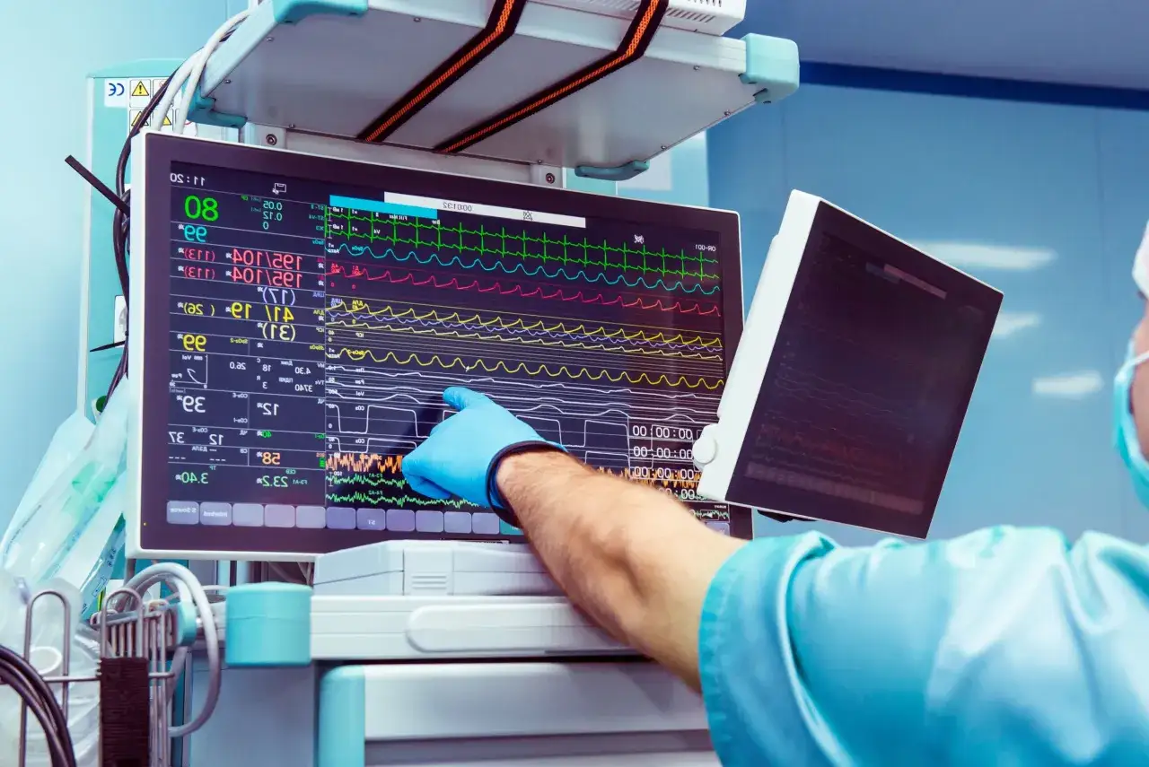 Lekarz w niebieskiej rękawiczce wskazuje na monitor parametrów życiowych, gdzie widoczne są krzywe EKG i inne wskaźniki. Może to być związane z podaniem leku, np. midazolam.