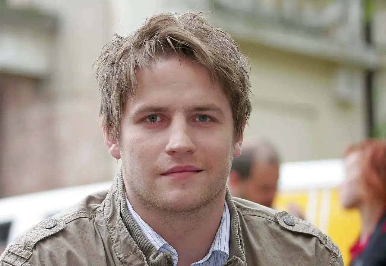 Kamil Maćkowiak w beżowej kurtce, z burzą blond włosów. Może właśnie myśli o swojej żonie.