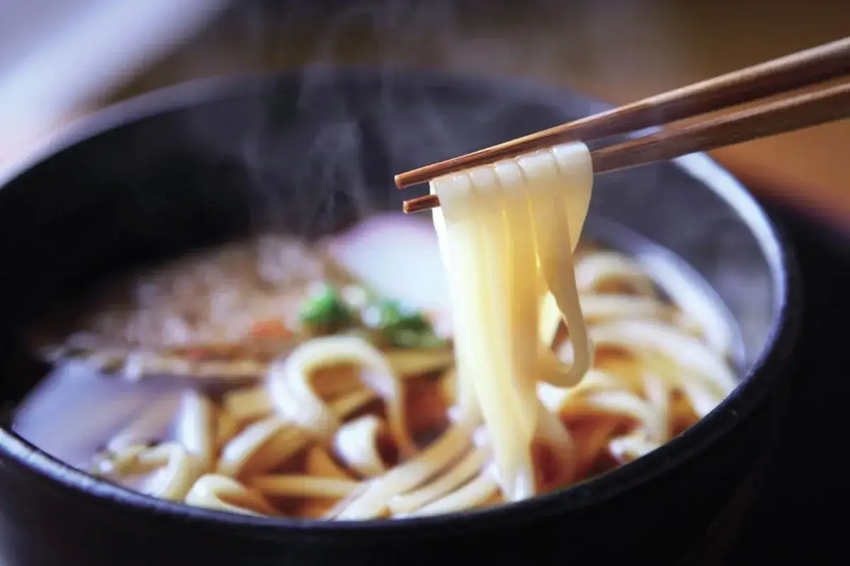 Para dowiedzieć się, jak gotować makaron udon, zobacz ten parujący talerz z grubym, żółtym makaronem podniesionym pałeczkami.