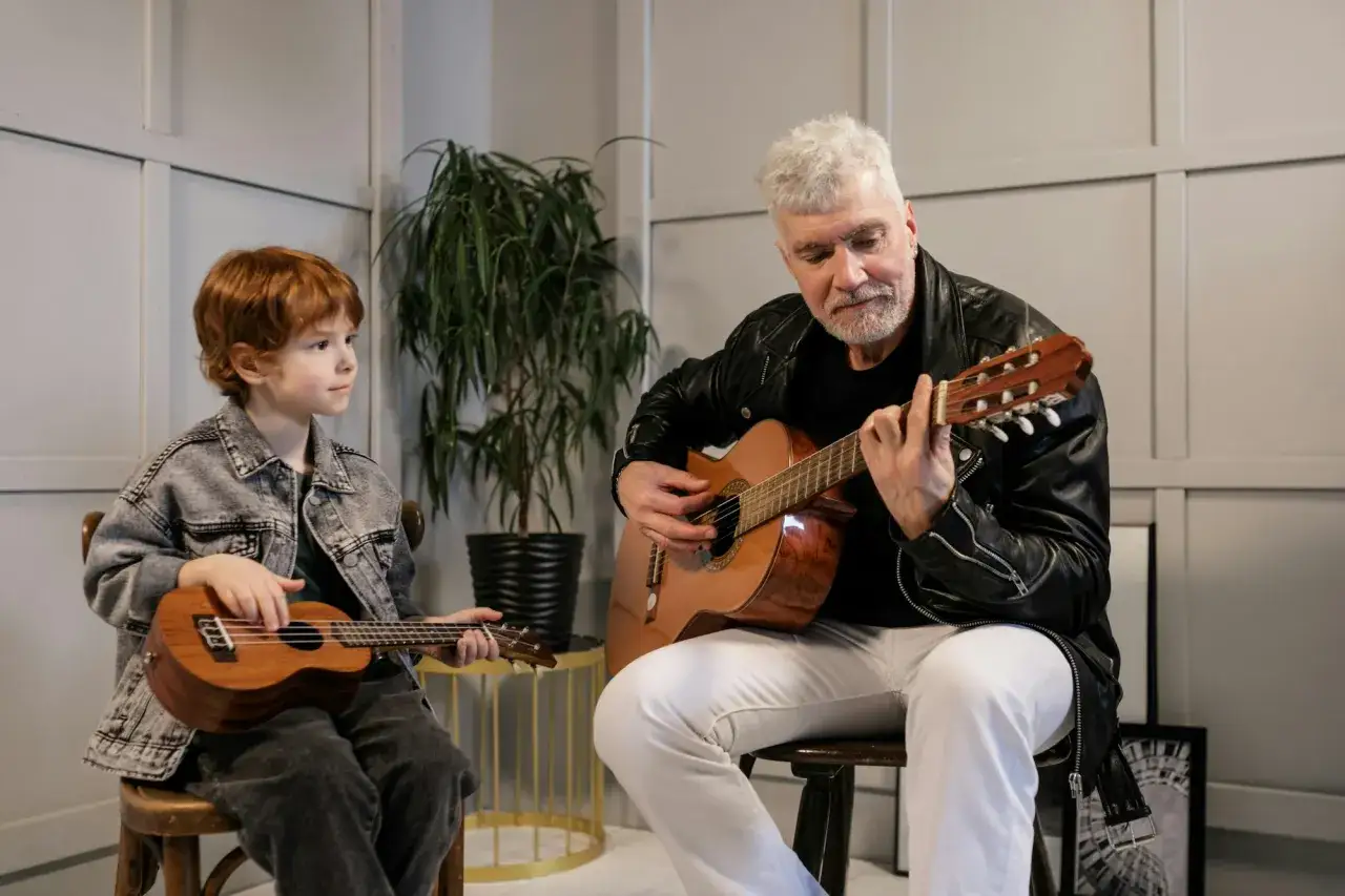 Dziadek uczy wnuka grać na gitarze w szkole muzycznej. Chłopiec trzyma ukulele, a starszy pan klasyczną gitarę.