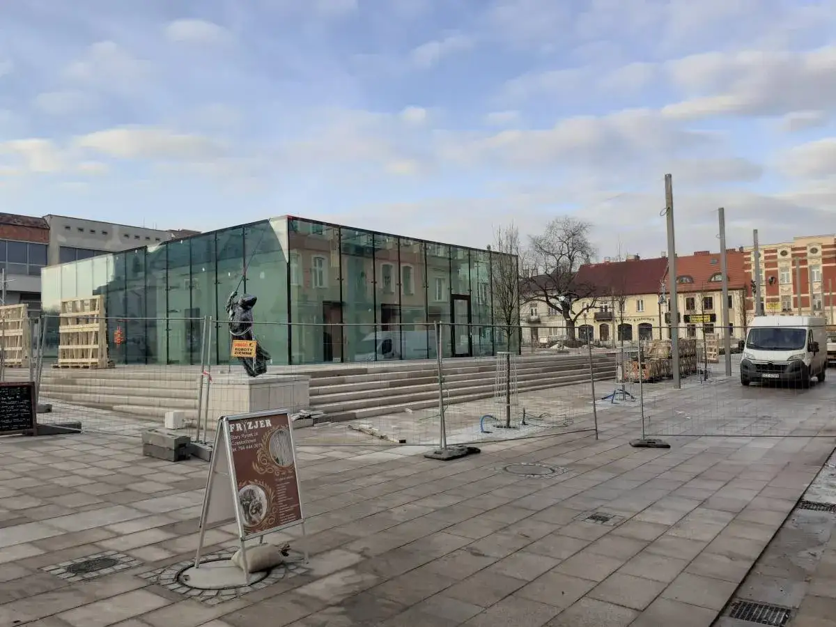 Nowoczesny budynek z przeszkloną fasadą na rynku Częstochowa. W pobliżu pomnik i tablica informacyjna.