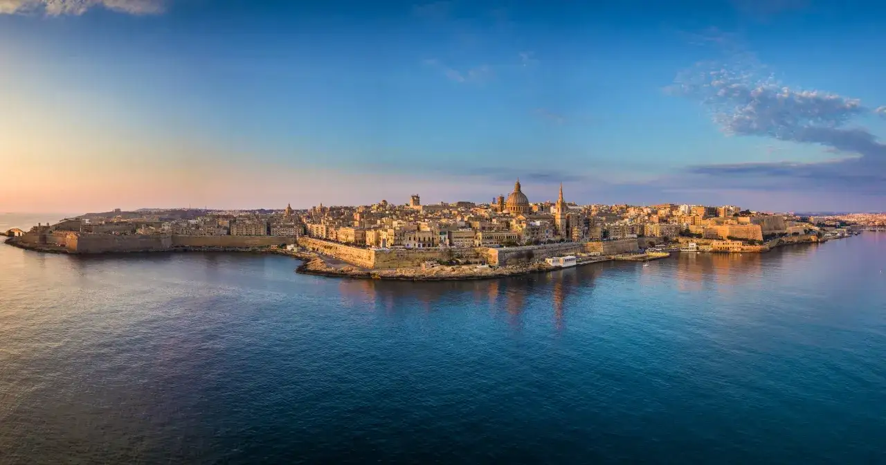 Malta w listopadzie zachwyca łagodną pogodą. Widok na Vallettę o wschodzie słońca, z jej fortyfikacjami i kopułami, odbijającymi się w spokojnym morzu.