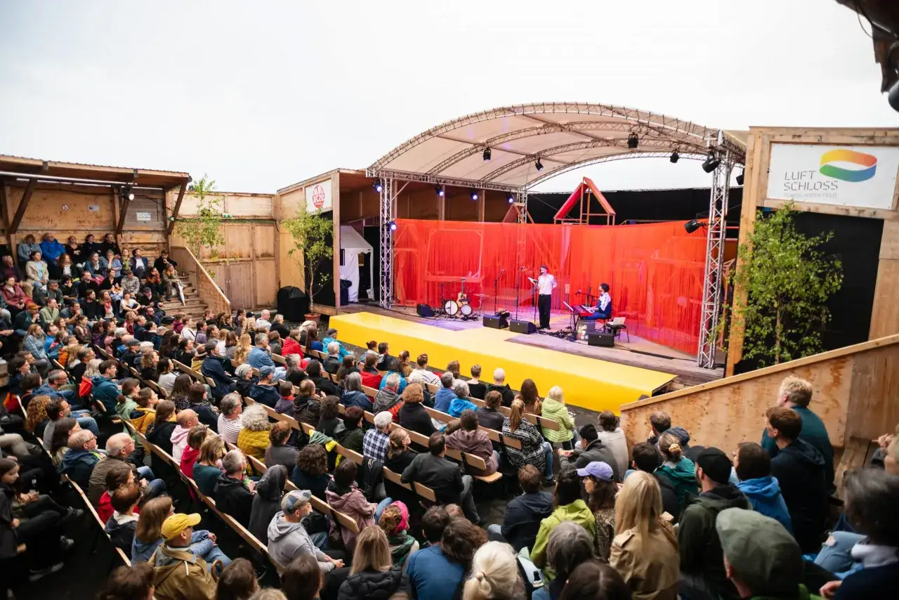 Publikum lauscht gespannt der Vorstellung im Luftschloss Tempelhofer Feld. Musiker und Sprecher auf der B&uuml;hne, rote Vorh&auml;nge und eine gelbe Rampe schaffen eine besondere Atmosph&auml;re.