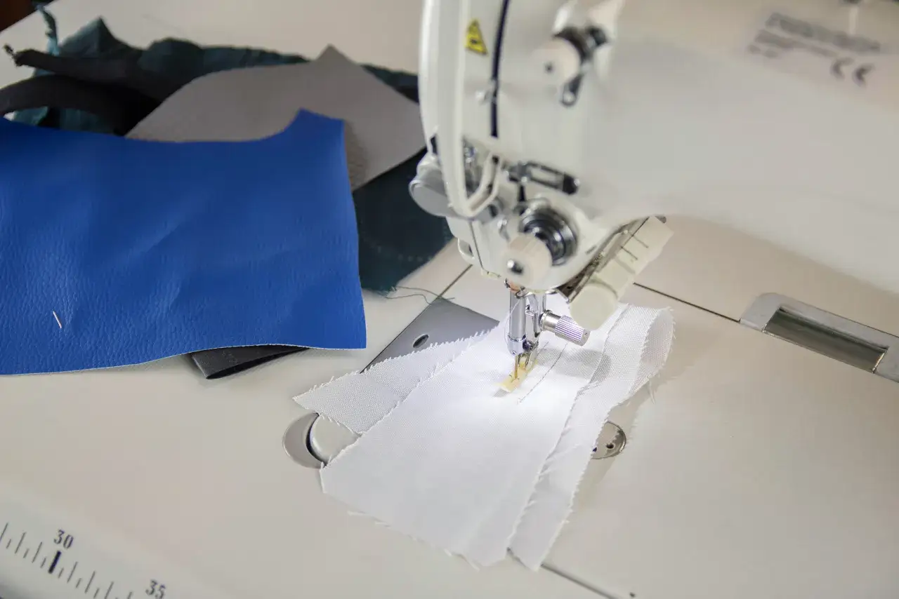 Maszyna do szycia wykonuje stebnowanie na białej tkaninie. Obok leżą próbki niebieskiej skóry i innych materiałów.