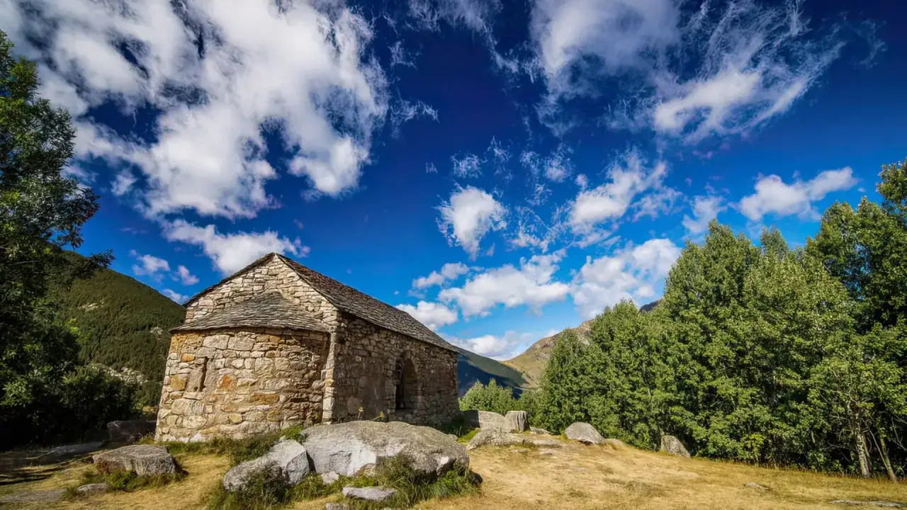 Pequeña ermita de piedra en las **rutas Vall de Boí**, rodeada de naturaleza y bajo un cielo azul con nubes algodonosas.