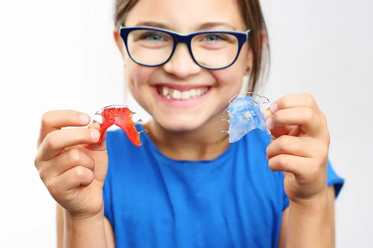 Bambina sorridente con occhiali mostra due espansori palato bambini, uno rosso e uno blu. Il costo è un fattore da considerare per la salute dentale.