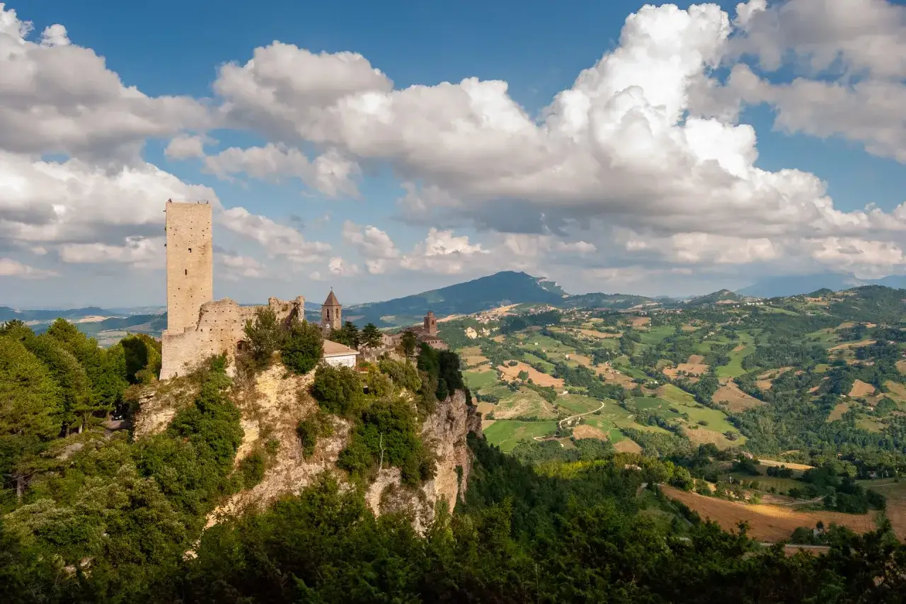 Rocche medievali svettano su colline verdi, un paesaggio tipico delle marche montagna. Nuvole bianche danzano nel cielo azzurro.