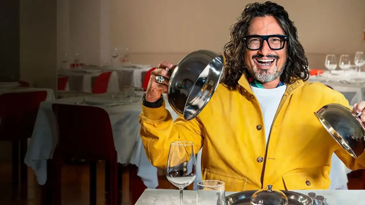 Chef con giacca gialla solleva un coperchio argentato in un ristorante, pronto per giudicare i piatti di 4 ristoranti.