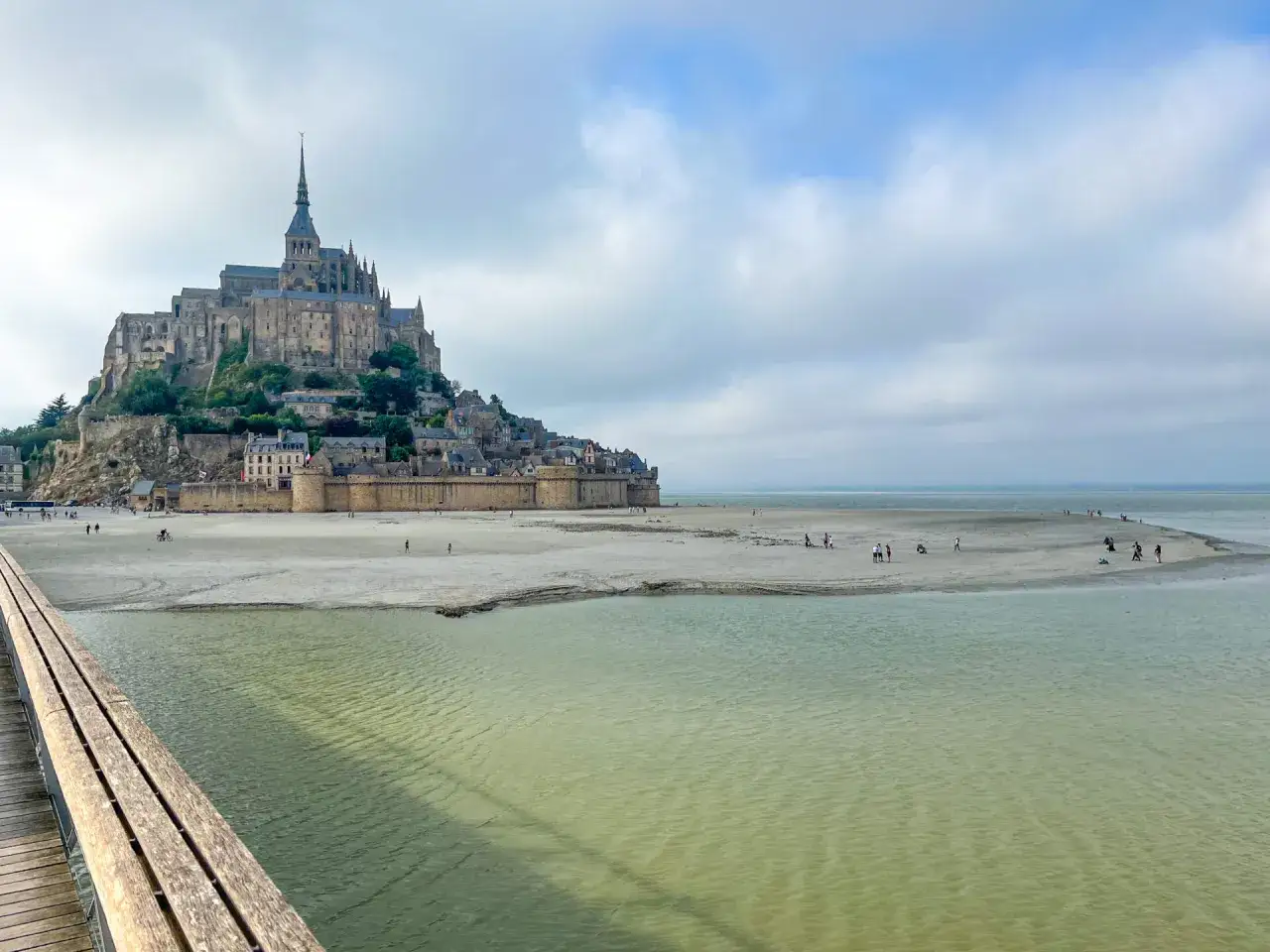 St. Michel, eine beeindruckende Abtei auf einer felsigen Insel, ragt &uuml;ber einer sandigen Bucht auf. Menschen spazieren am Ufer entlang.