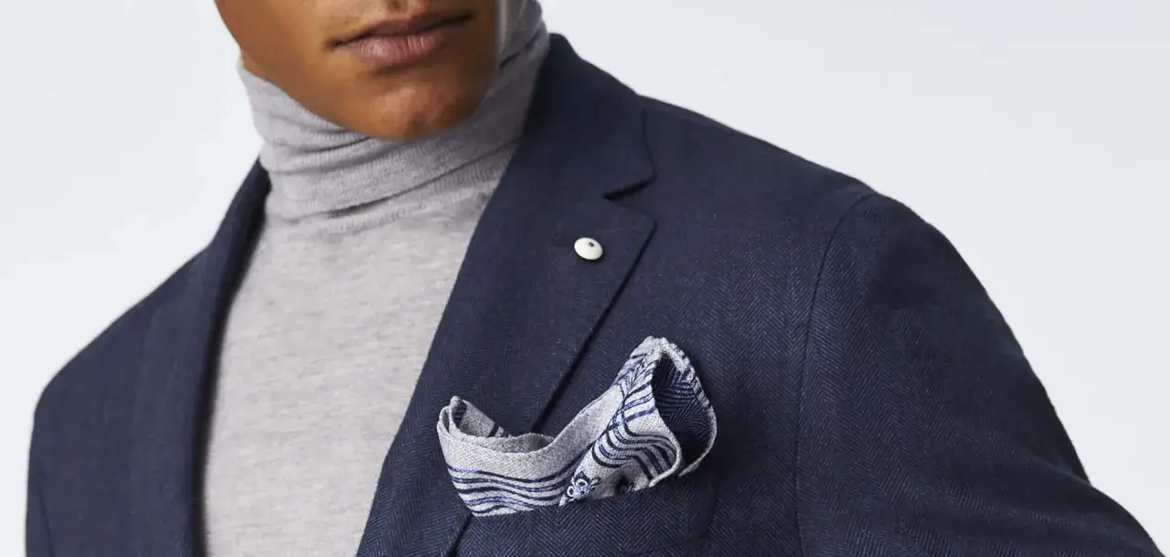 Un uomo indossa un blazer blu scuro con un dolcevita grigio e un fazzoletto con un motivo blu e bianco. Il fazzoletto, un accessorio di stile, aggiunge un tocco di eleganza.