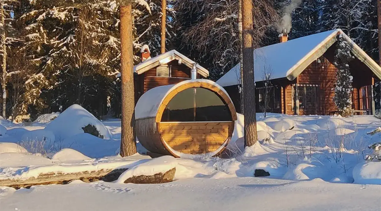 En tunna bastu i snöig skogsmiljö i Sverige. Röken stiger från skorstenen på ett närliggande hus.