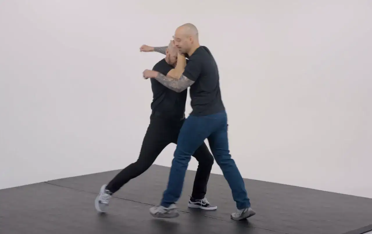 Two men practice boxing form, one in black pants and the other in blue jeans, demonstrating a close-quarters grapple.