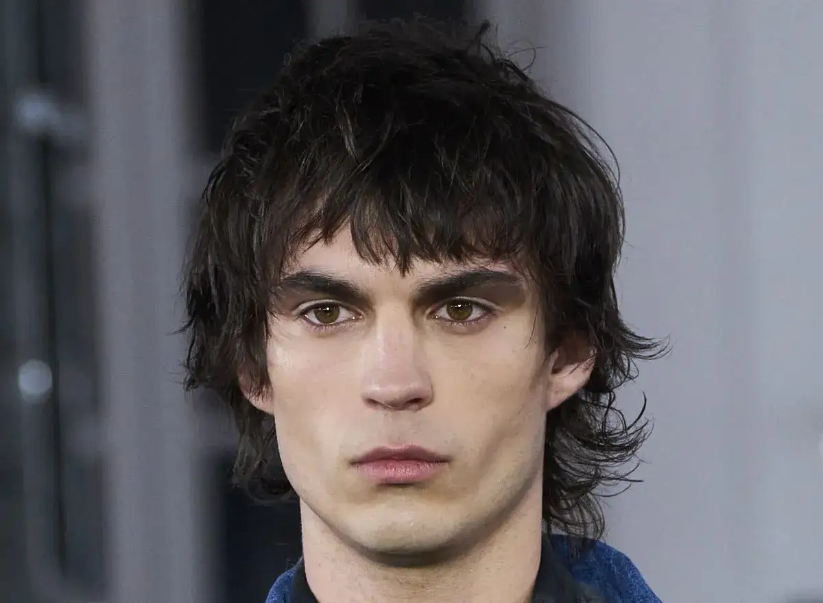 Primer plano de un hombre joven con un corte de pelo mullet desordenado y ojos penetrantes.