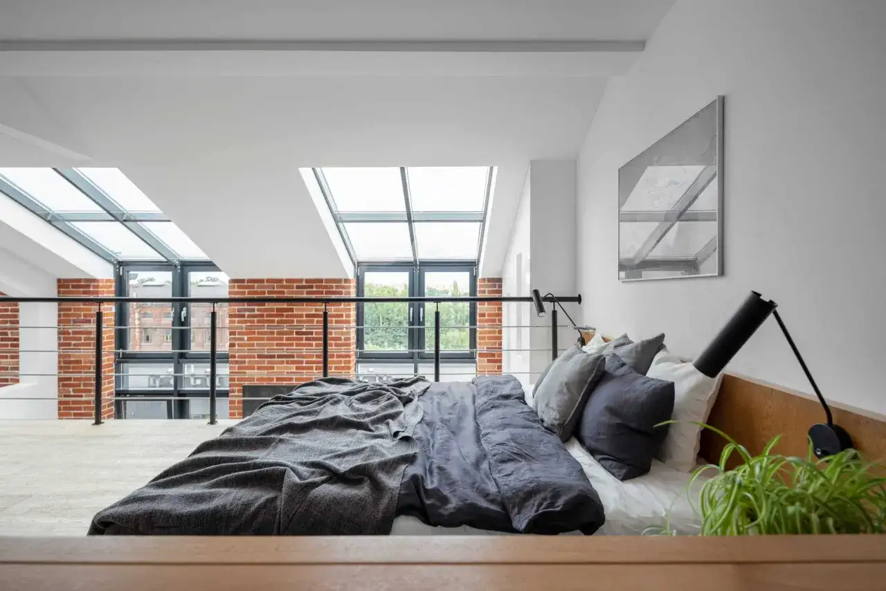 Lit douillet dans une chambre mezzanine avec vue sur des briques apparentes et un ciel lumineux par les fenêtres de toit.