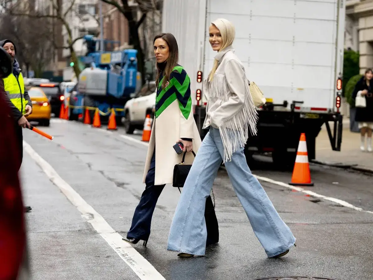 Dos mujeres cruzan la calle, una con un abrigo llamativo y la otra con un pantalón flare de mezclilla y chaqueta de flecos.