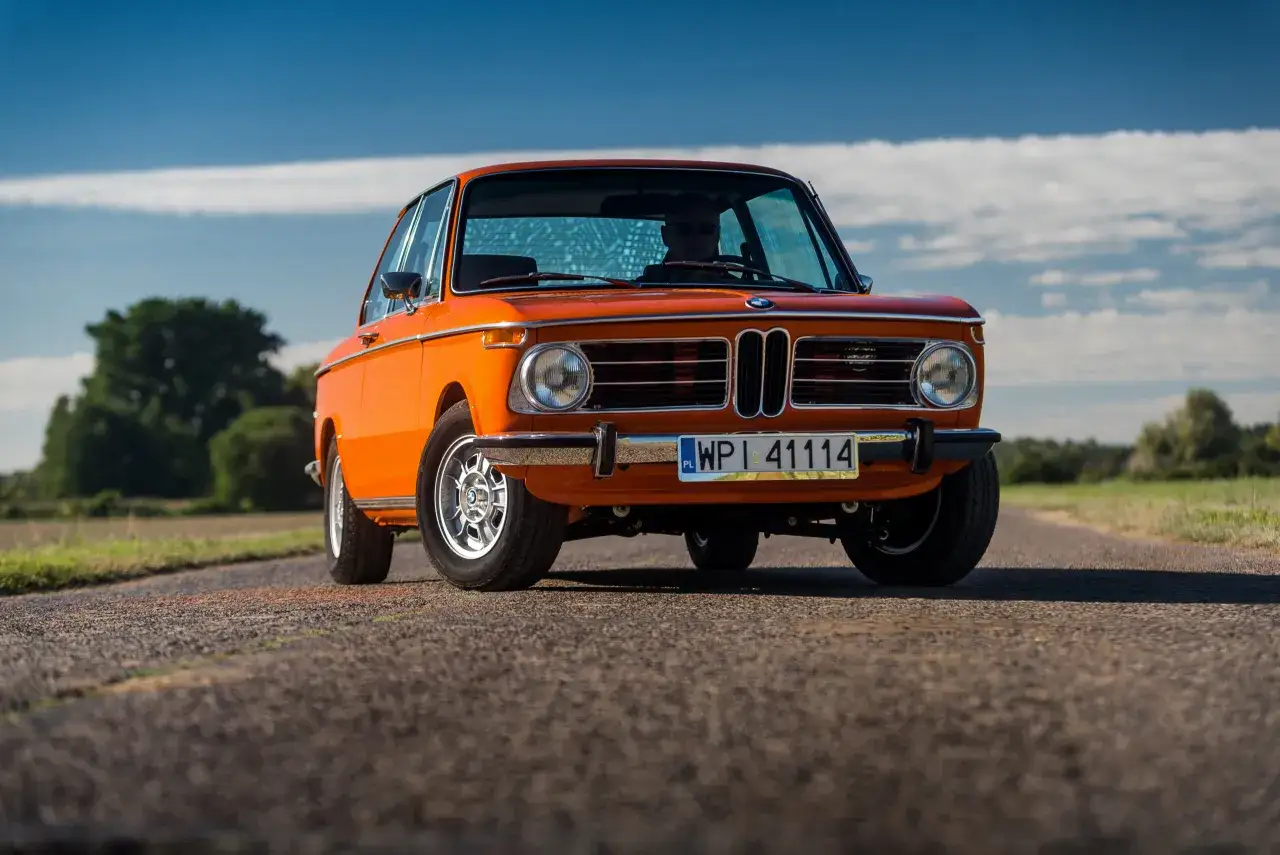 Pomarańczowe BMW 2002 na pustej drodze, z kierowcą w okularach przeciwsłonecznych za kierownicą.