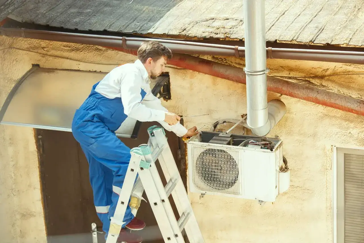Serwisant na drabinie naprawia klimatyzację. Dowiedz się, ile kosztuje serwis klimatyzacji w domu.