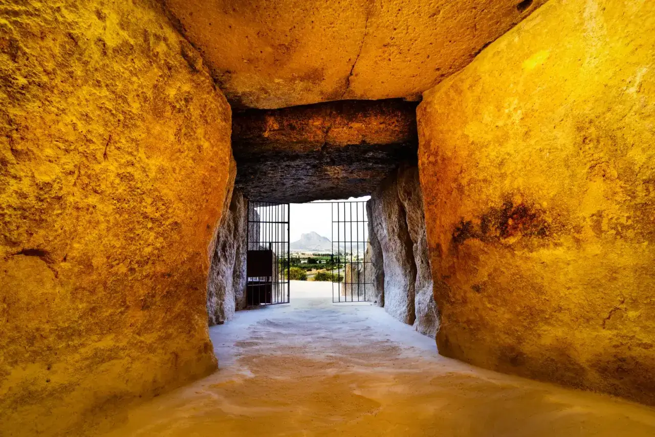 Interior del dolmen de Menga, con sus enormes losas de piedra y una reja que da al exterior, donde se vislumbra un paisaje montañoso.