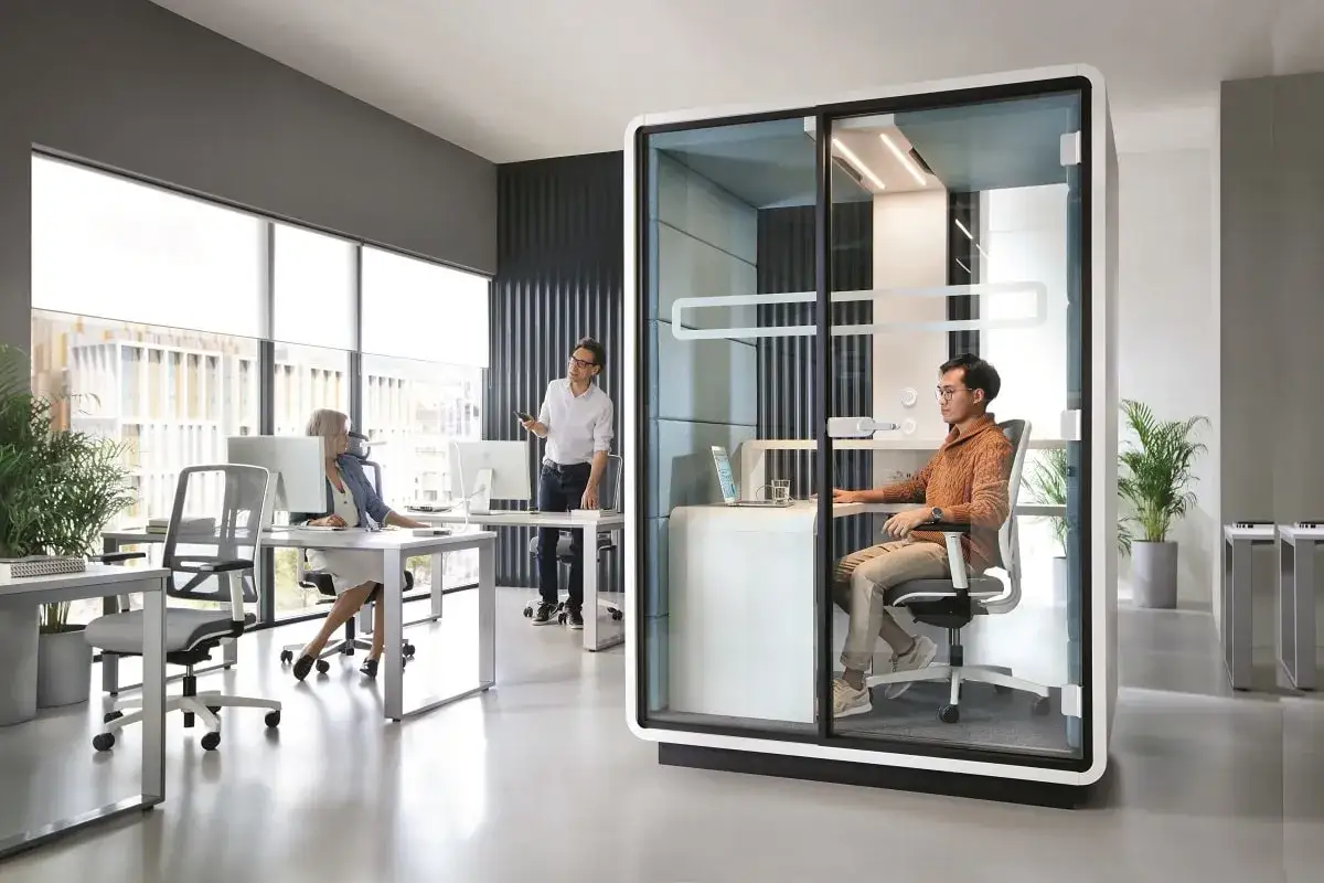 Bureau moderne avec un espace ouvert. Un homme travaille dans une cabine vitrée, tandis que deux collègues discutent près des fenêtres.