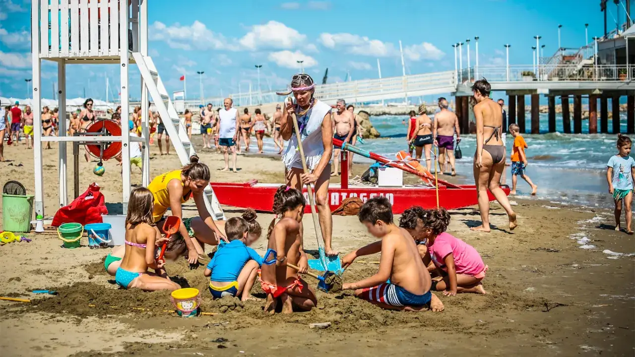 Bambini giocano sulla sabbia a Gabicce Mare centro, mentre bagnanti si godono il mare e la spiaggia affollata.