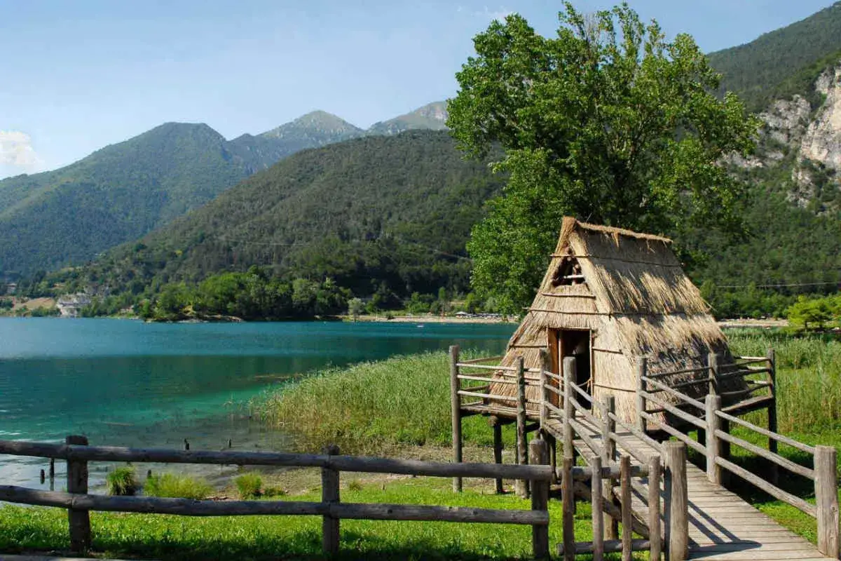 Capanna di paglia su palafitte a Bardolino, con lago turchese e montagne verdi sullo sfondo.