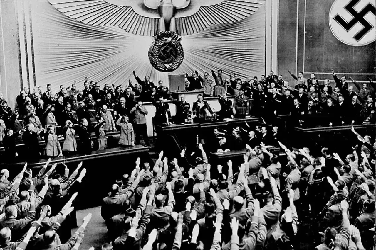 Stor folksamling med uppstr&auml;ckta armar och nazistiska symboler. En man i uniform, m&ouml;jligen Hitler, st&aring;r p&aring; en upph&ouml;jd plats.