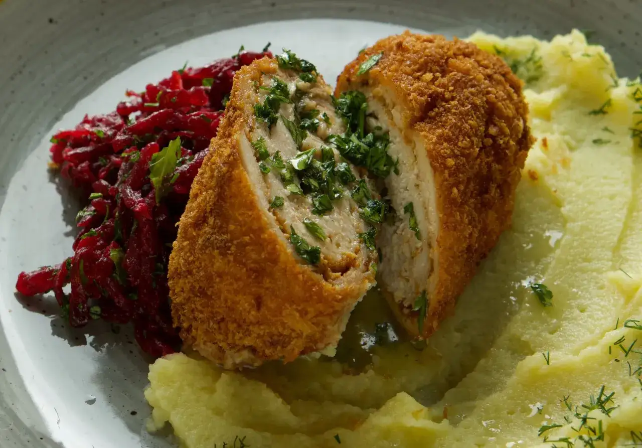 Chrupiący kotlet po kijowsku, przekrojony, odsłaniający soczyste wnętrze z ziołami, podany z pur&eacute;e ziemniaczanym i buraczkami.