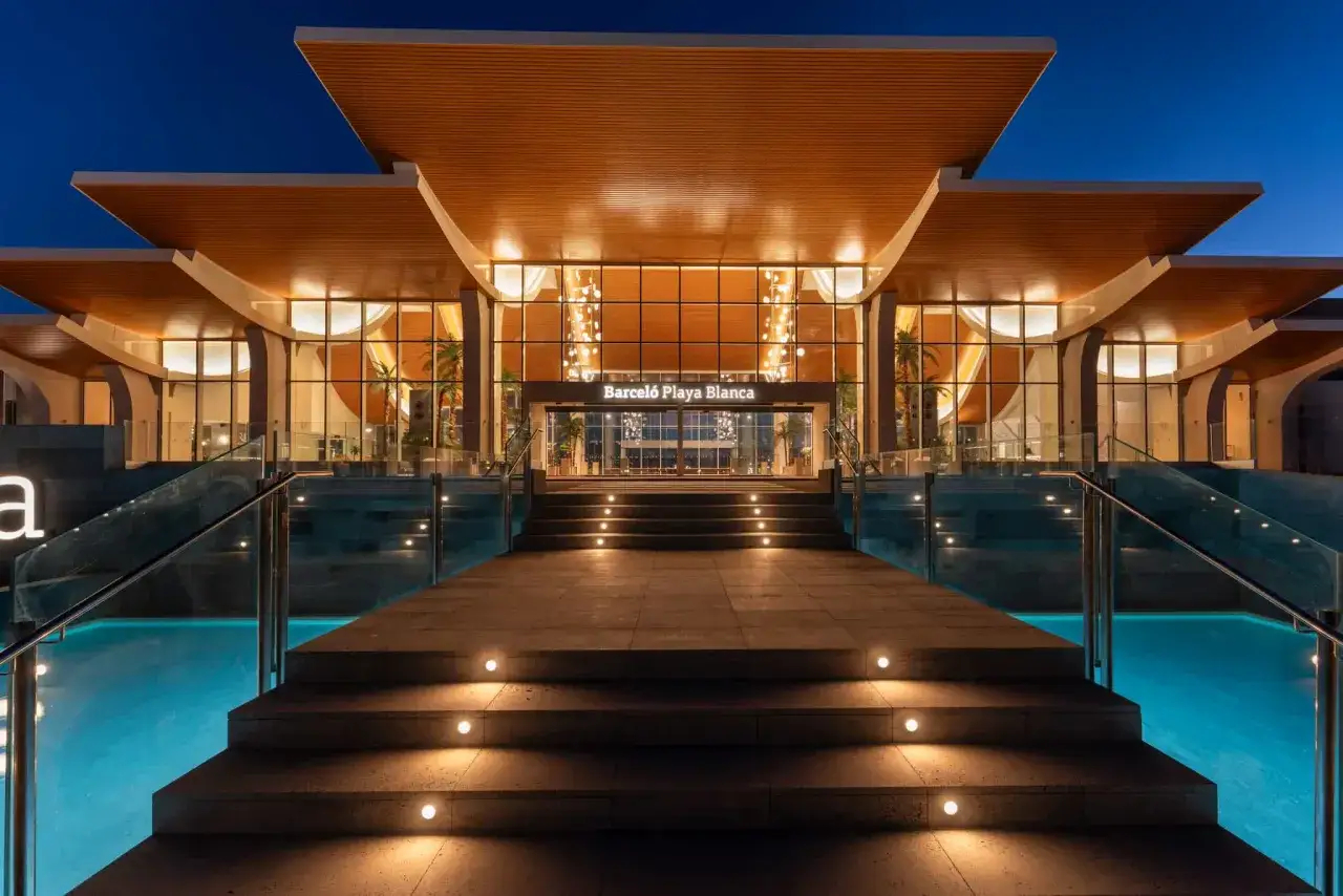 Eleganckie wejście do Barceló Playa Blanca oświetlone nocą. Schody prowadzą do przeszklonego lobby, obok którego lśni basen.