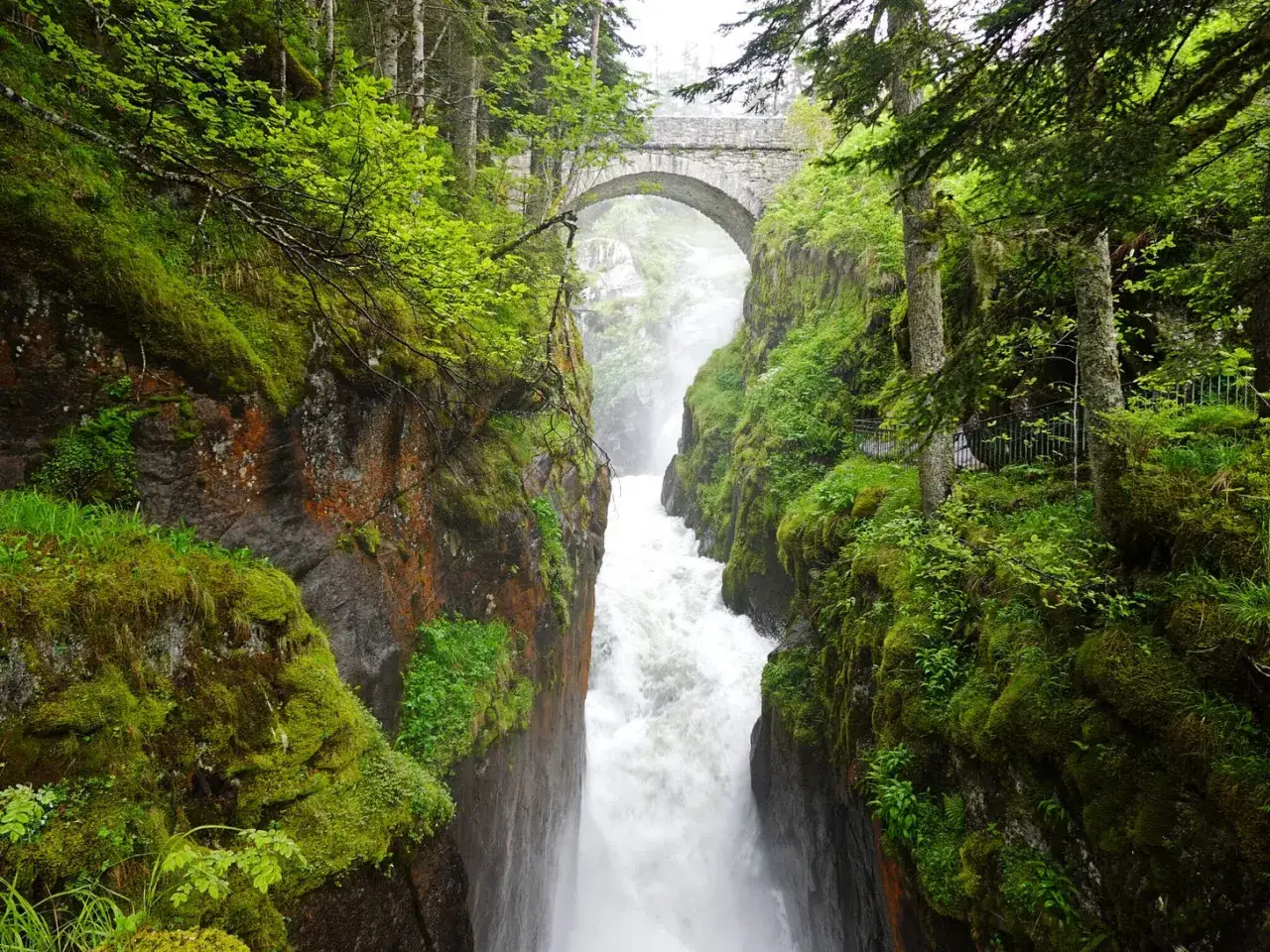 Un puente de piedra arqueado cruza un ca&ntilde;&oacute;n verde exuberante, con una cascada rugiente cayendo entre rocas cubiertas de musgo. El **pont de espagne** se alza majestuoso sobre la naturaleza salvaje.