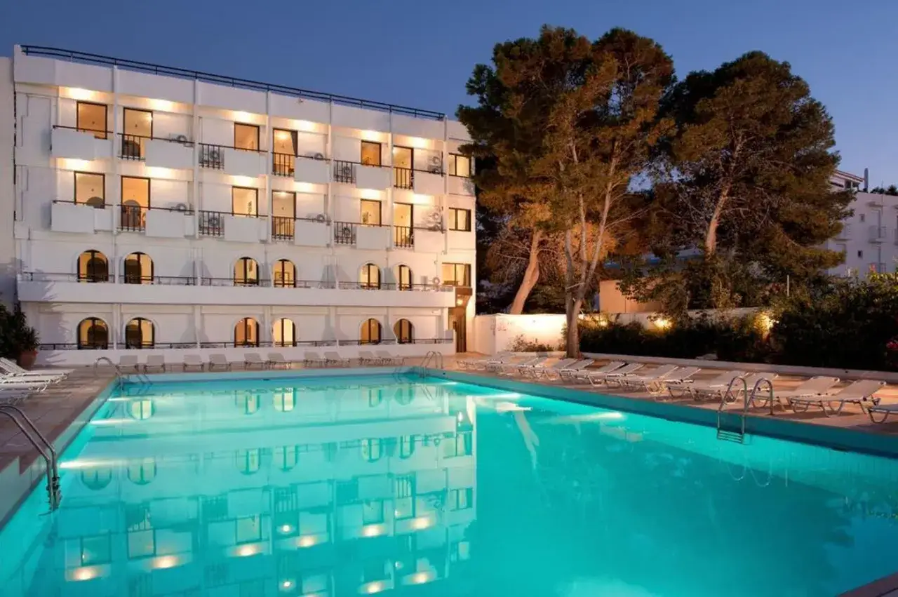 Basen hotelowy wieczorem, odbijający światła. W tle biały budynek z balkonami. Wiele osób szuka Heronissos Hotel opinie, by sprawdzić ten obiekt.
