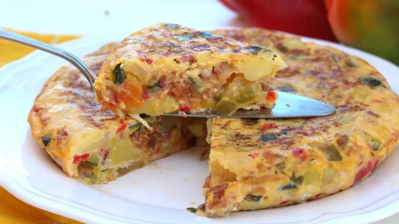 Una porción de tortilla paisana, jugosa y llena de verduras como pimientos y calabacín, se levanta con una espátula.