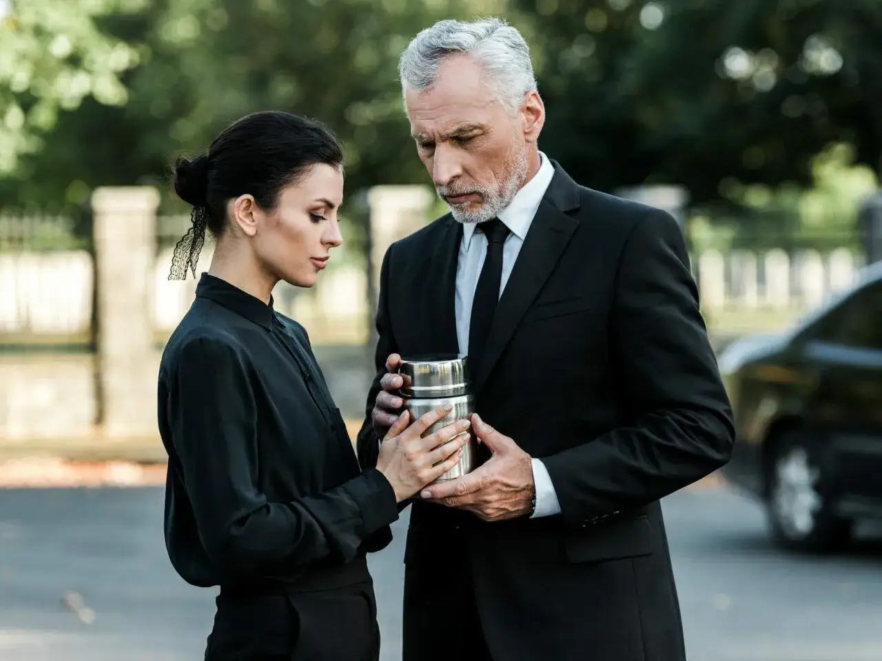 Mężczyzna w garniturze i kobieta w czarnej sukience trzymają urnę. Elegancki ubiór męski na pogrzebie latem.