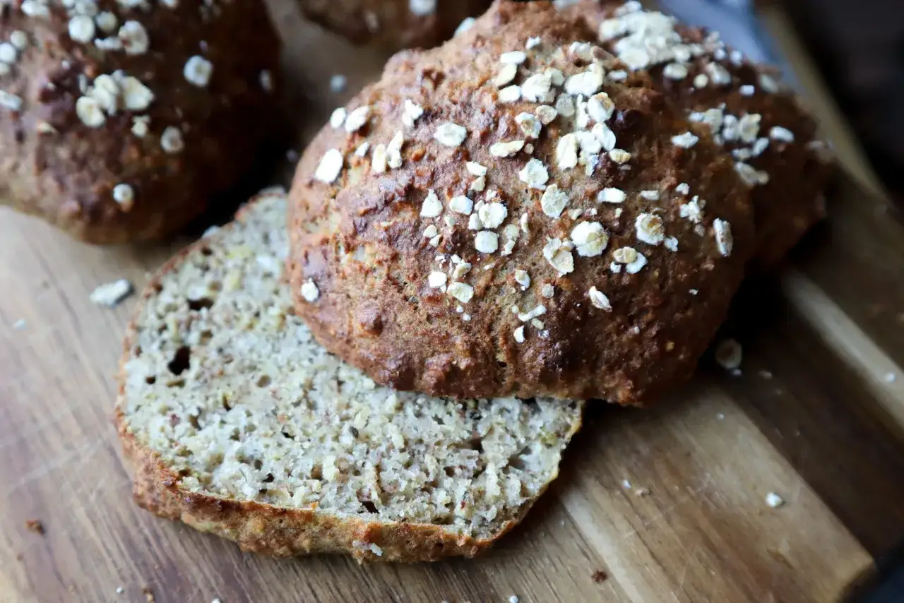 Nybakade glutenfria frallor med havregryn p&aring; en tr&auml;sk&auml;rbr&auml;da. En fralla &auml;r uppskuren och visar ett saftigt inre.
