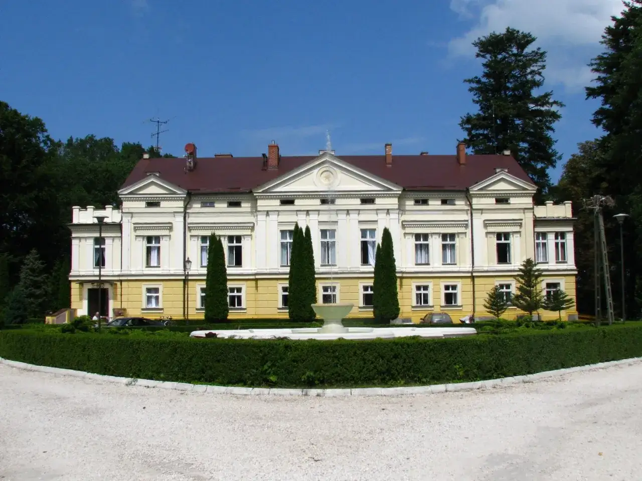 Pałac w Sasinie, okazała budowla z ż&oacute;łtą fasadą i czerwoną dach&oacute;wką, otoczony zielenią i fontanną.