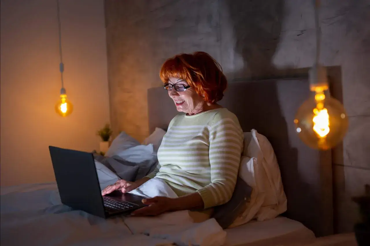 Uśmiechnięta starsza kobieta z rudymi włosami i okularami korzysta z laptopa w łóżku. To dowód, że **ageizm** jest nieuzasadniony – wiek to tylko liczba.