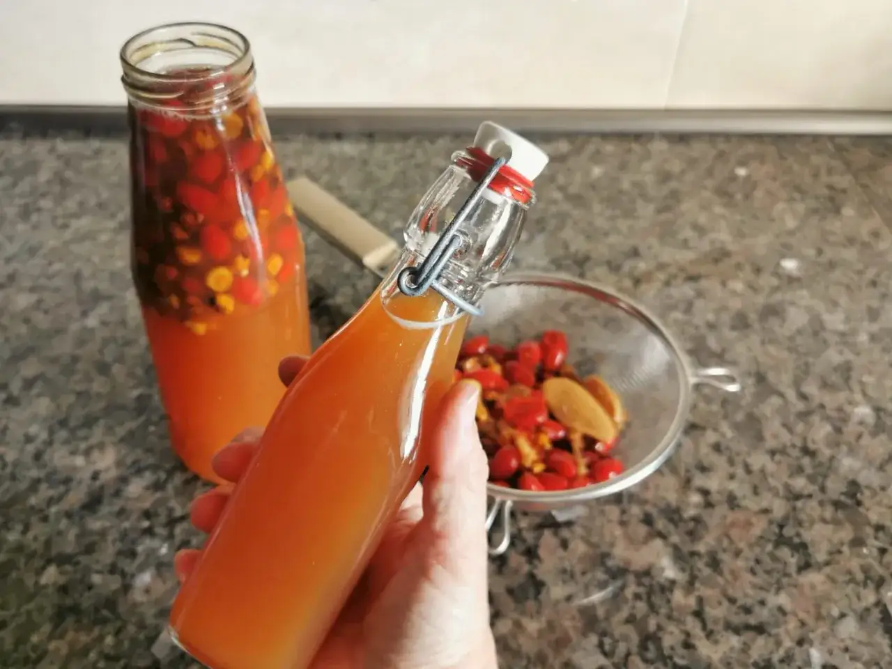 Eine Hand hält eine Flasche mit einem bernsteinfarbenen Getränk. Im Hintergrund eine weitere Flasche und ein Sieb mit Hagebutten. Ein selbstgemachter Oxymel Hagebutte.