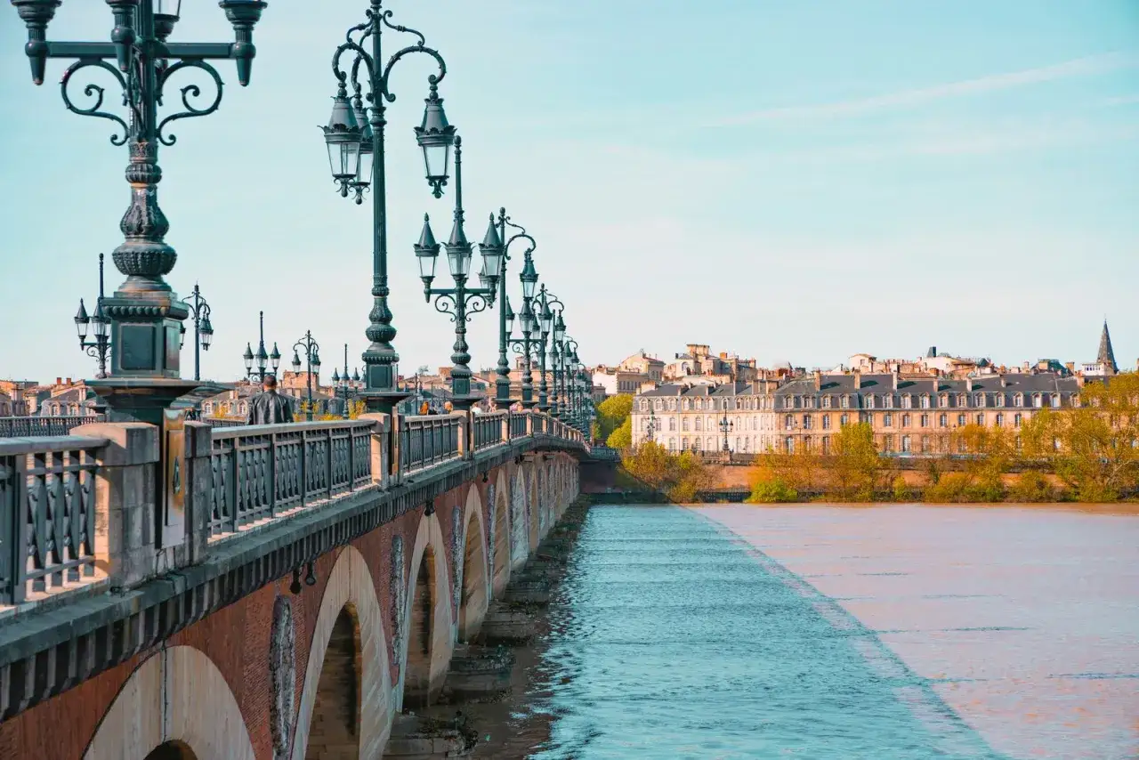 A man walks across a picturesque bridge in Bordeaux, with ornate lampposts and historic buildings lining the river. Is Bordeaux worth visiting? Absolutely!