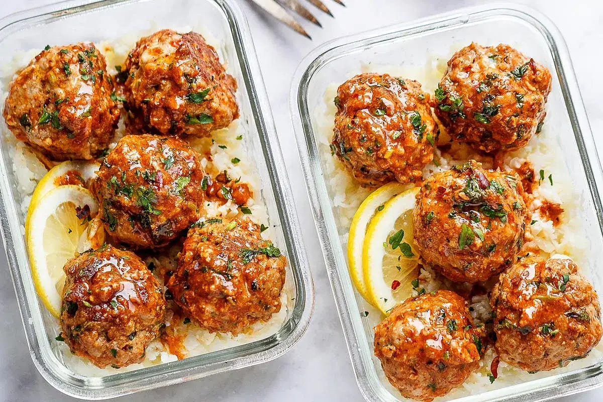 Zwei Behälter mit Hackbällchen in Sauce auf Reis, garniert mit Zitrone und Petersilie. Perfekt für ein schnelles Mittagessen warm.