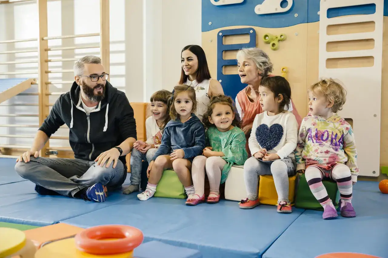 Grupa dzieci i dorosłych siedzi na macie w sali przedszkolnej. Dzieci słuchają opowieści, zastanawiając się, ile trwa przedszkole.