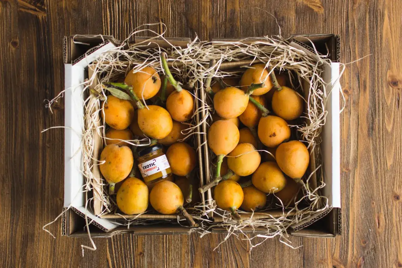 Caja llena de nísperos frescos, destacando sus propiedades. Un frasco de mermelada de kumquat acompaña la fruta.