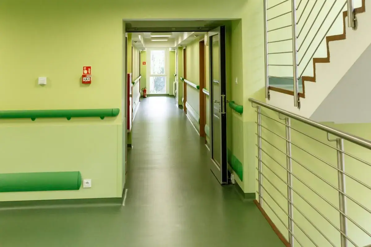 Jasny korytarz z zielonymi poręczami i podłogą. Wygląda jak dom emeryta wojskowego, gdzie każdy szczegół zapewnia bezpieczeństwo i komfort.