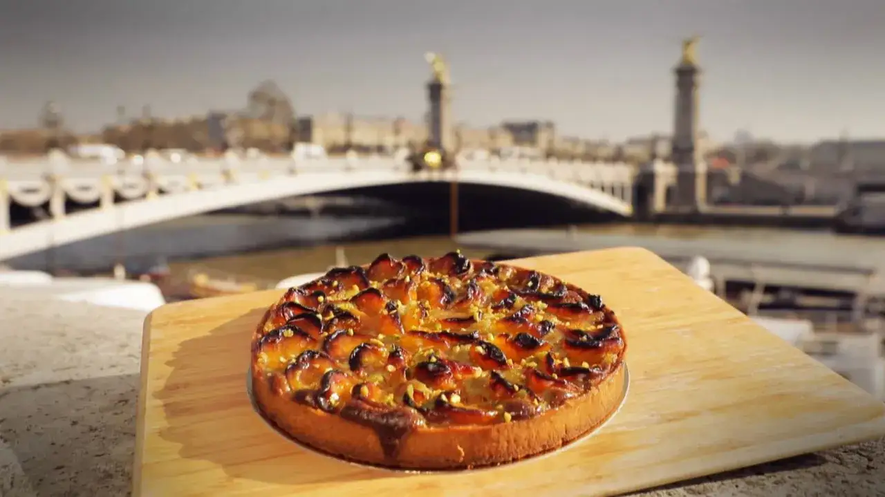 Tarte abricot pistache dorée, posée sur une planche en bois, avec un pont parisien en arrière-plan.