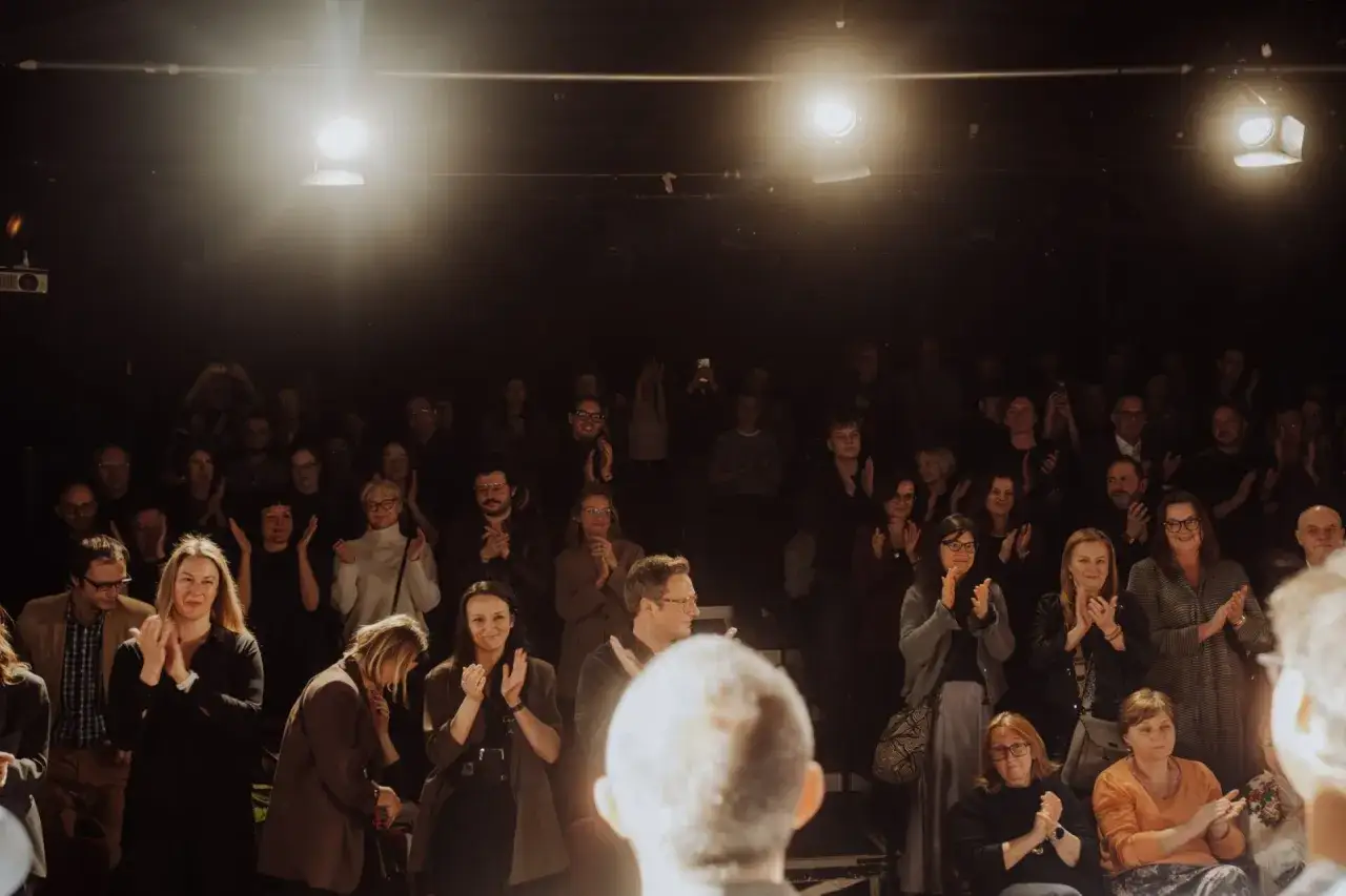 Widownia klaszcze po udanej **performance**. Ludzie w różnym wieku, oświetleni reflektorami, wyrażają swoje uznanie.