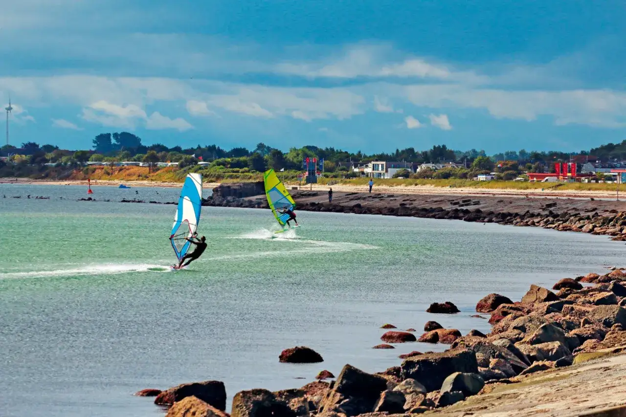 Zwei Windsurfer gleiten über das Wasser. Im Hintergrund eine Küstenlandschaft mit Häusern und Bäumen. Ein typischer Tag für Windsurfen Deutschland.