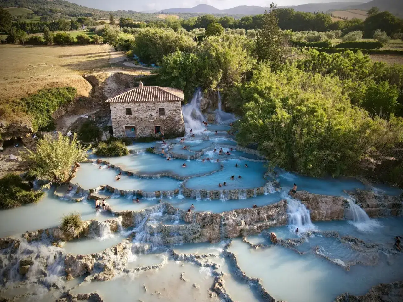 Cascades d'eau thermale bleue à Rapolano Terme, avec une vieille bâtisse en pierre et des baigneurs profitant de la détente.