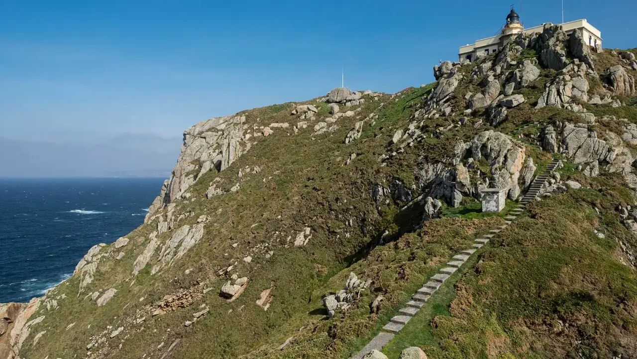 Escaleras de piedra ascienden por la ladera verde y rocosa del cabo Priorino, coronada por un faro blanco.