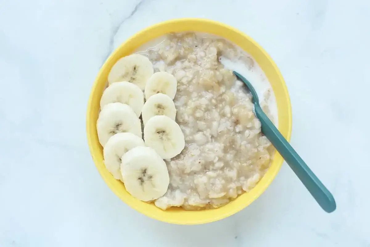 Una ciotola gialla piena di fiocchi d'avena cremosi con fette di banana disposte elegantemente. Un cucchiaio verde è immerso nel porridge.