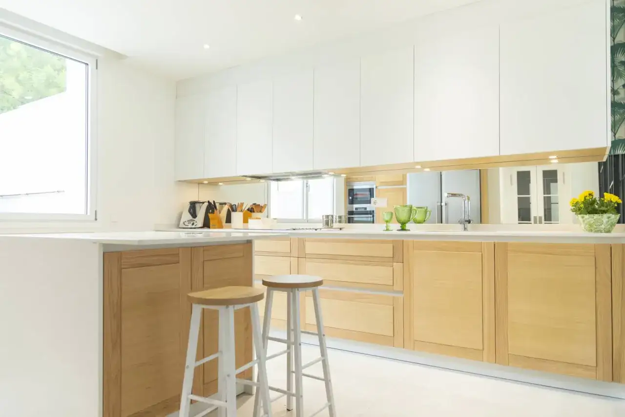 Moderna cocina blanca y madera clara, con isla central y taburetes. Un espacio luminoso y funcional.