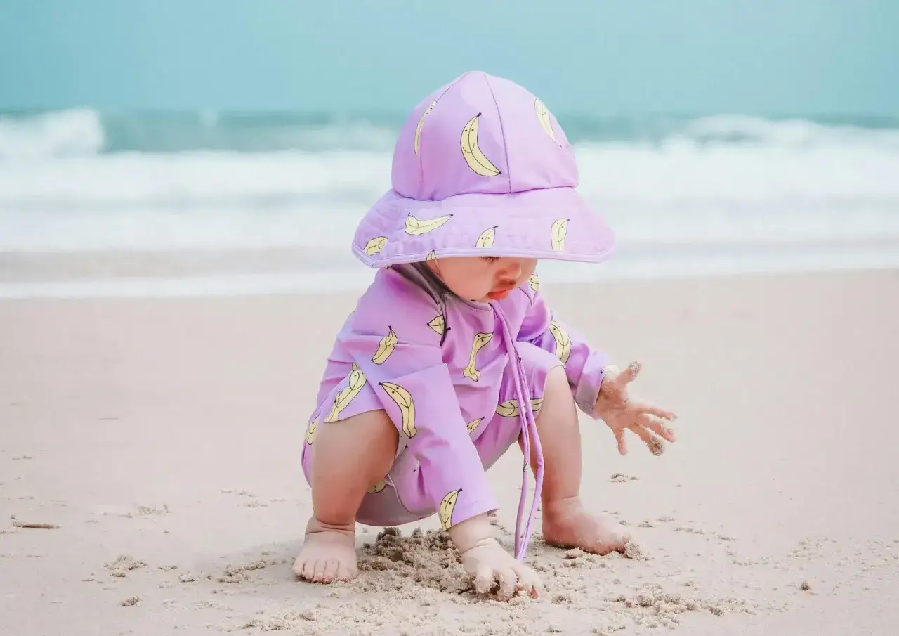 Niemowlę w fioletowym stroju w banany na plaży. Po kąpieli można wyjść na dwór z niemowlakiem, gdy jest ciepło i słonecznie.