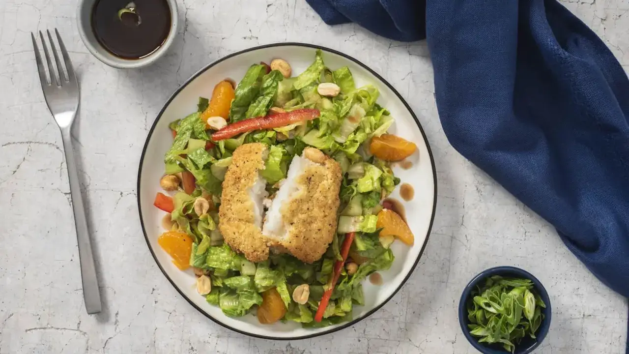 Une salade fraîche et colorée avec un poisson pané croustillant, des quartiers d'orange, des poivrons rouges et des cacahuètes.
